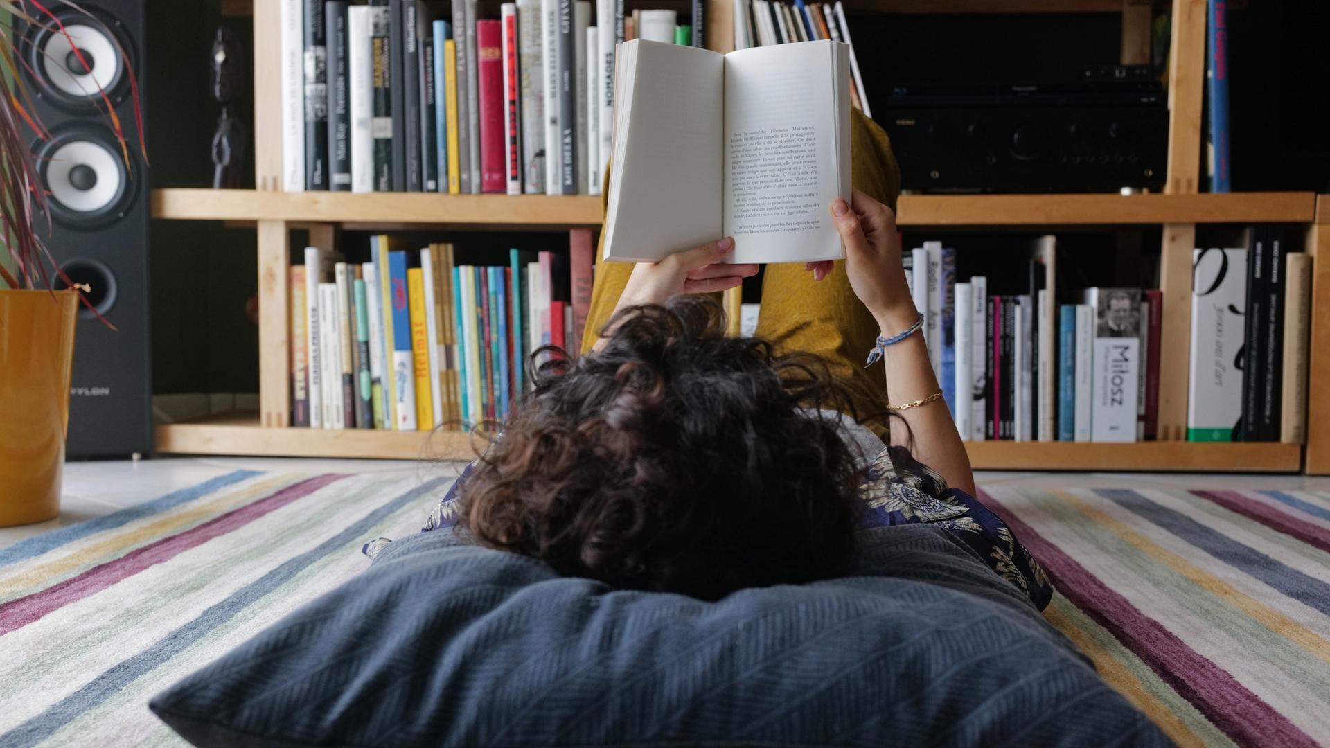 Eine junge Person liegt auf dem Boden und liest ein Buch. Der Kopf ist nur von hinten zu sehen und liegt auf einem großen grauen Kissen. 