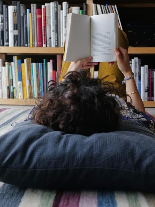 Eine junge Person liegt auf dem Boden und liest ein Buch. Der Kopf ist nur von hinten zu sehen und liegt auf einem großen grauen Kissen. 