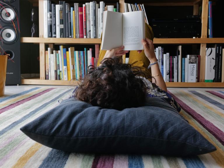 Eine junge Person liegt auf dem Boden und liest ein Buch. Der Kopf ist nur von hinten zu sehen und liegt auf einem großen grauen Kissen. 