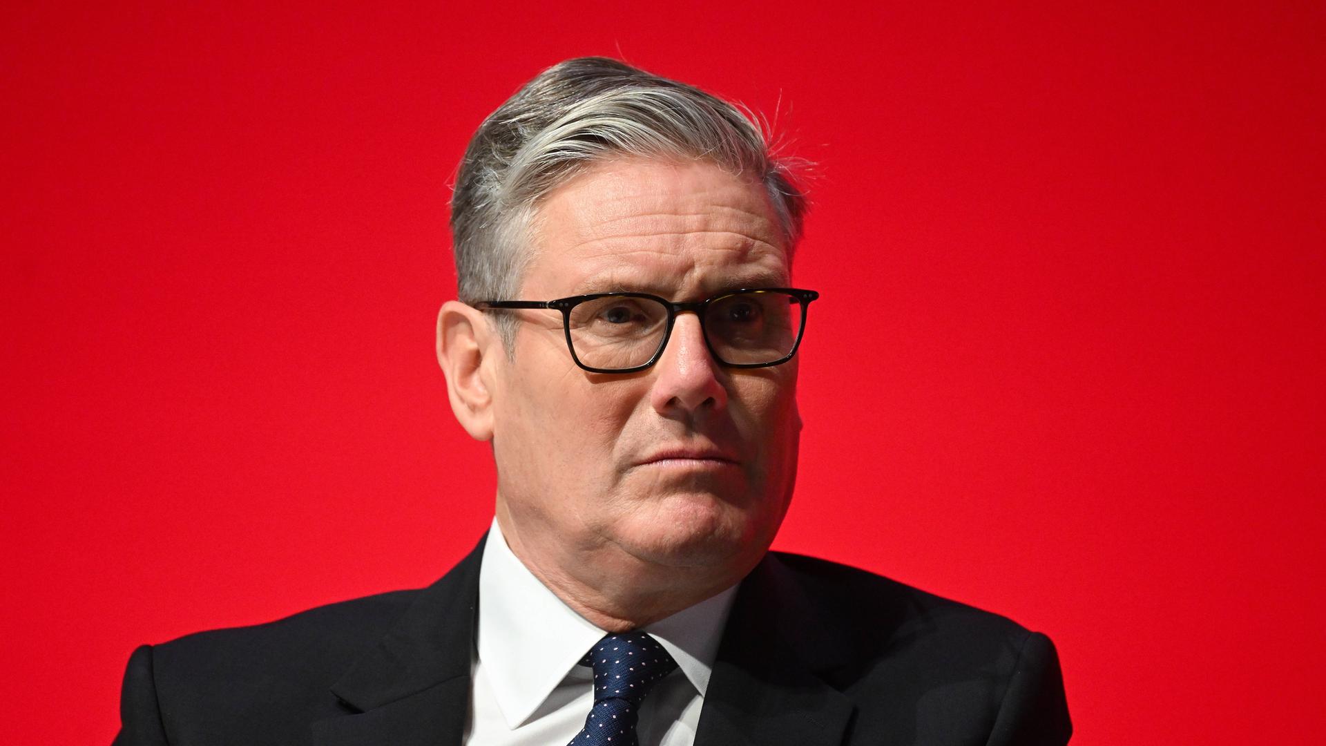 Keir Starmer in einem schwarzen Anzug vor einer roten Wand
