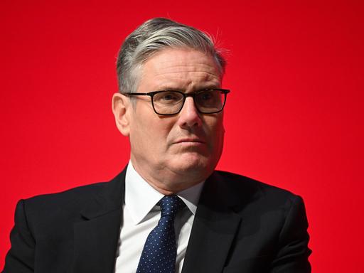 Keir Starmer in einem schwarzen Anzug vor einer roten Wand