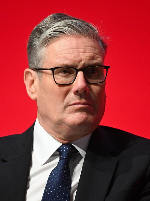 Keir Starmer in einem schwarzen Anzug vor einer roten Wand