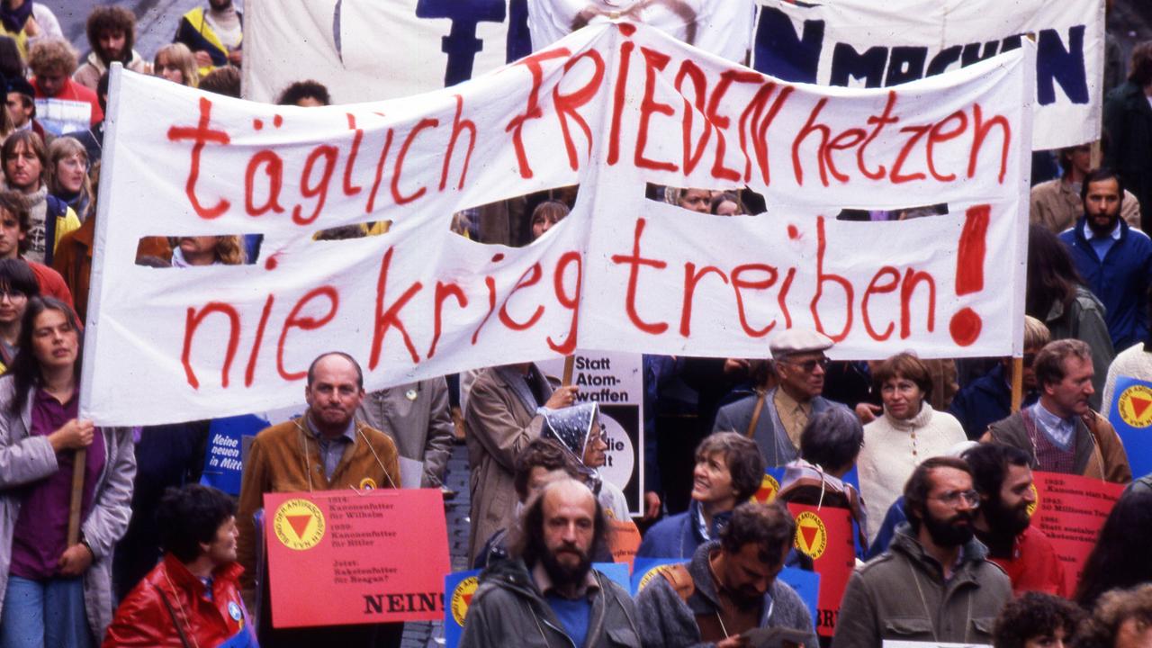 Foto einer Demonstration auf der unter anderem Banner mit der Aufschrift "Täglich Frieden hetzen - nie Krieg treiben" von Menschen gehalten wird. Davor sind weitere Personen mit Schildern zu sehen, die die Aufschrift "Keine Atomraketen" tragen.