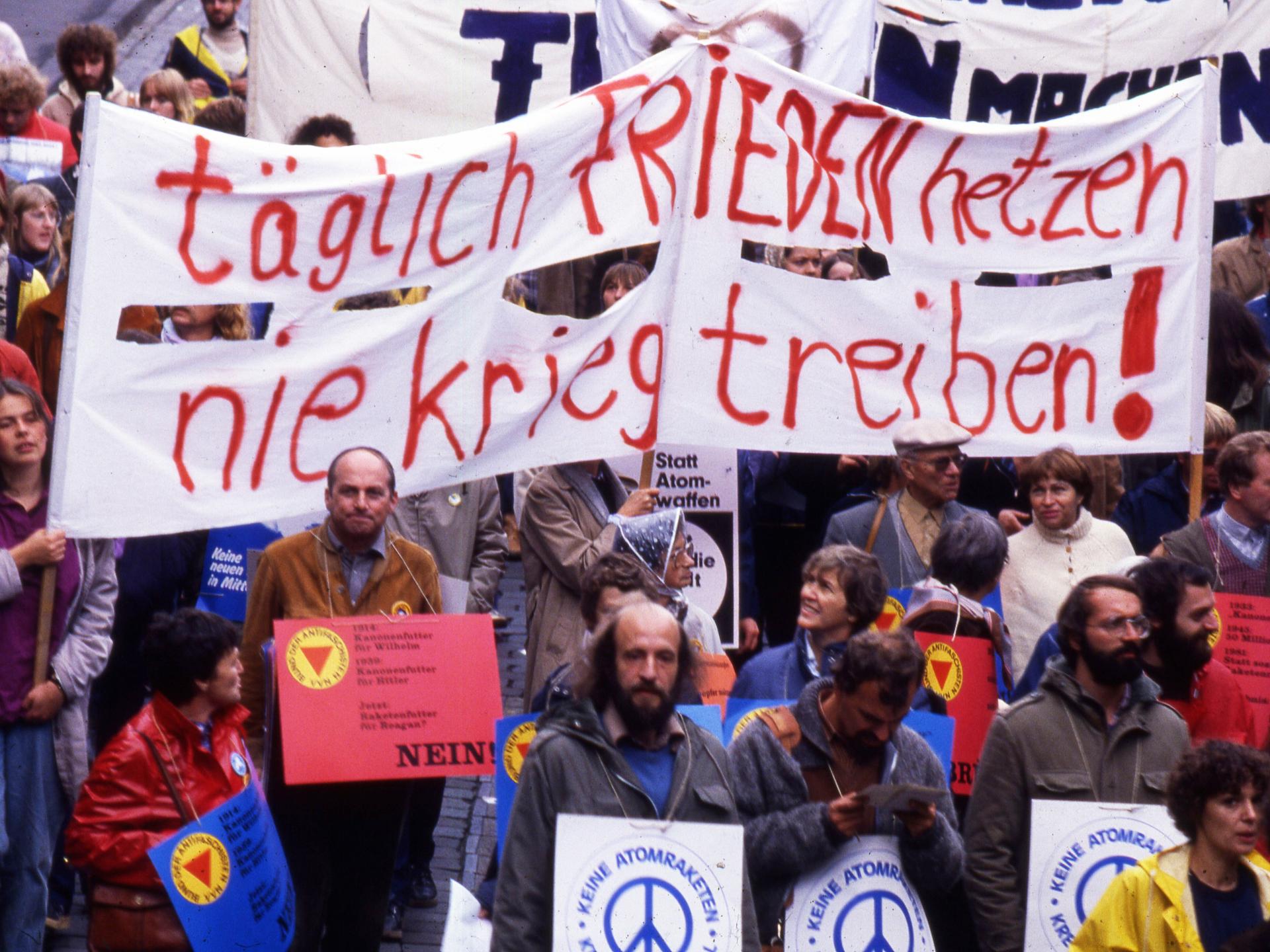 Foto einer Demonstration auf der unter anderem Banner mit der Aufschrift "Täglich Frieden hetzen - nie Krieg treiben" von Menschen gehalten wird. Davor sind weitere Personen mit Schildern zu sehen, die die Aufschrift "Keine Atomraketen" tragen.