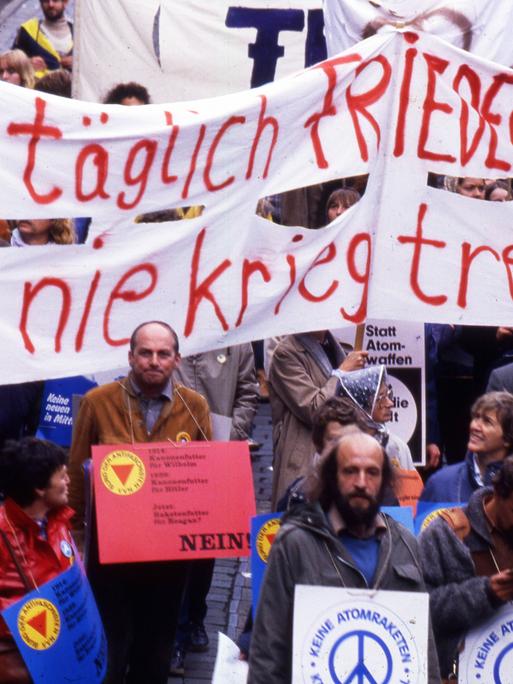 Foto einer Demonstration auf der unter anderem Banner mit der Aufschrift "Täglich Frieden hetzen - nie Krieg treiben" von Menschen gehalten wird. Davor sind weitere Personen mit Schildern zu sehen, die die Aufschrift "Keine Atomraketen" tragen.