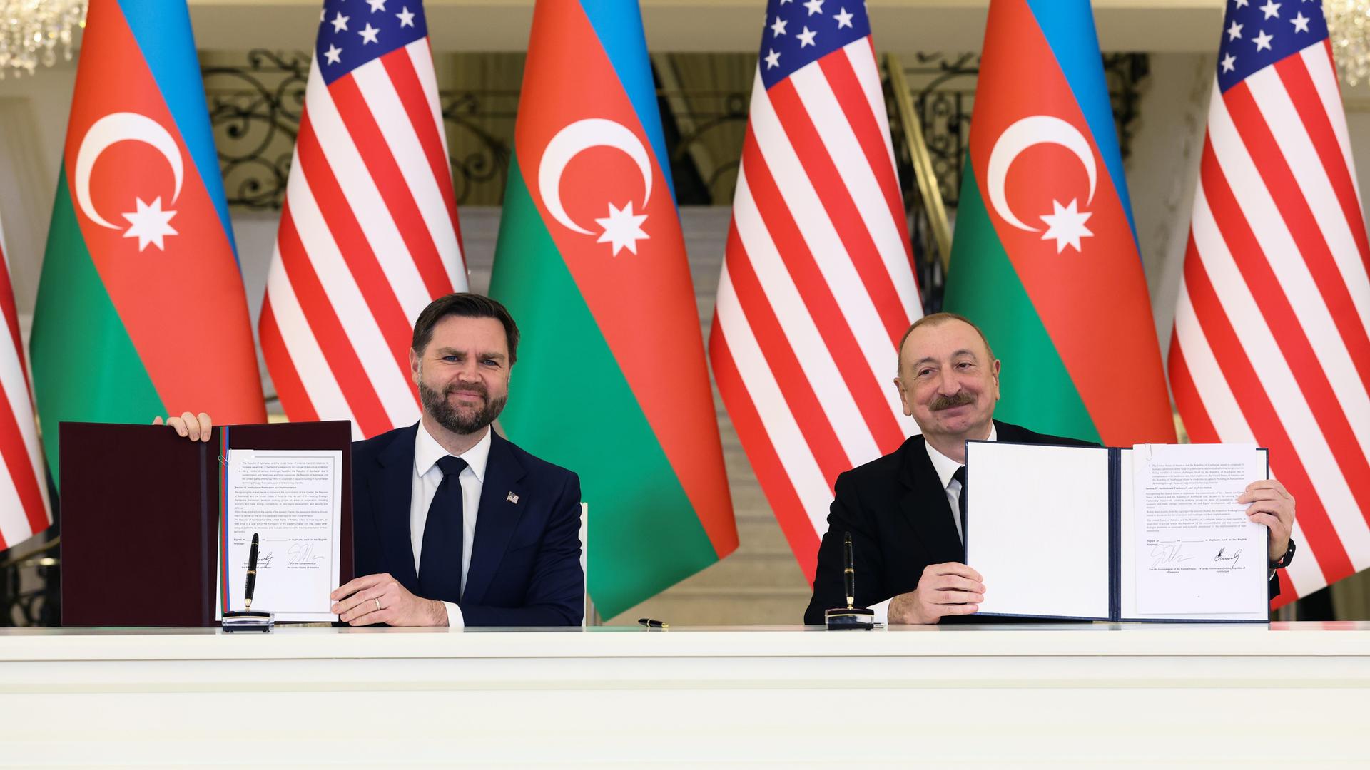 US-Vizepräsident JD Vance (l) und der aserbaidschanische Präsidenten Ilham Alijew sitzen vor aserbaidschanischen und US-amerikanischen Flaggen. Sie halten unterzeichnete Verträge in ihren Händen.