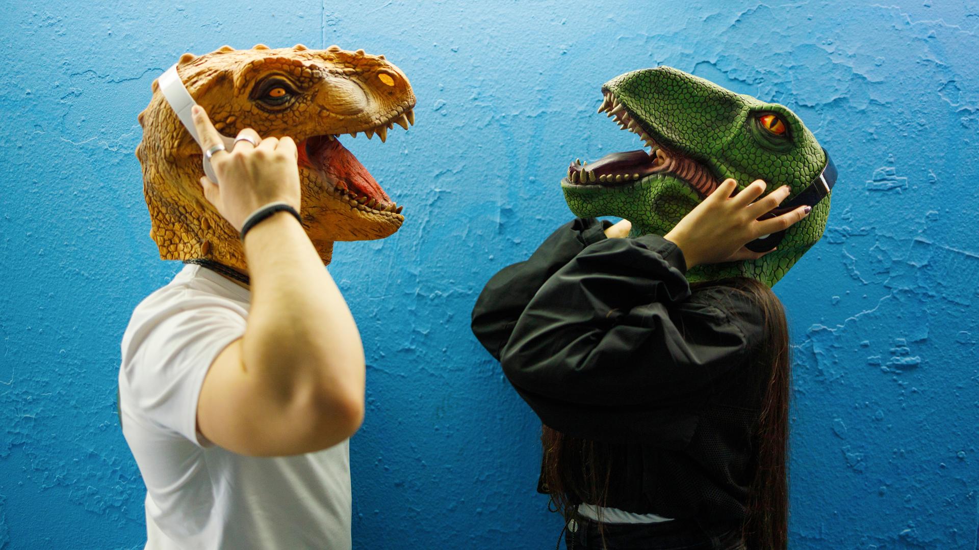 Zwei Menschen mit Dinosauriermaske tragen Kopfhörer und hören Podcasts.