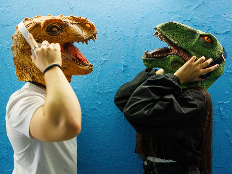 Zwei Menschen mit Dinosauriermaske tragen Kopfhörer und hören Podcasts.