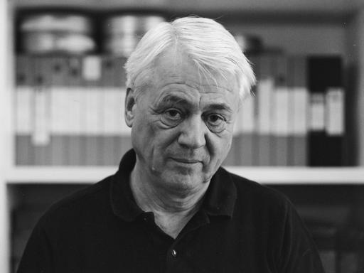 Schwarzweiße Porträtaufnahme zeigt Alexander Kluge vor einem Bücherregal mit Filmrollen im Jahr 1997