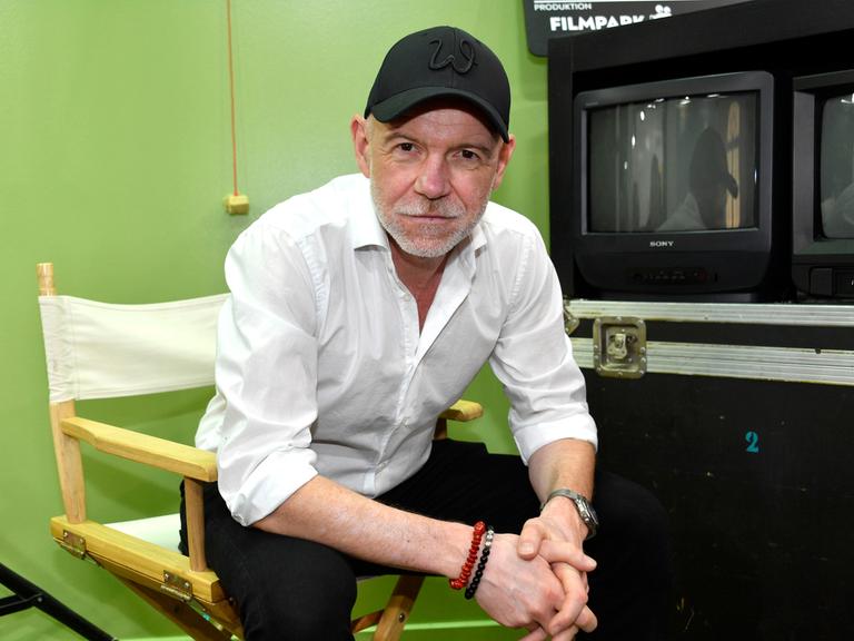 Dietmar Wunder sitzt in einem Studio im Filmpark Babelsberg auf einem Regiestuhl und schaut freundlich in die Kamera