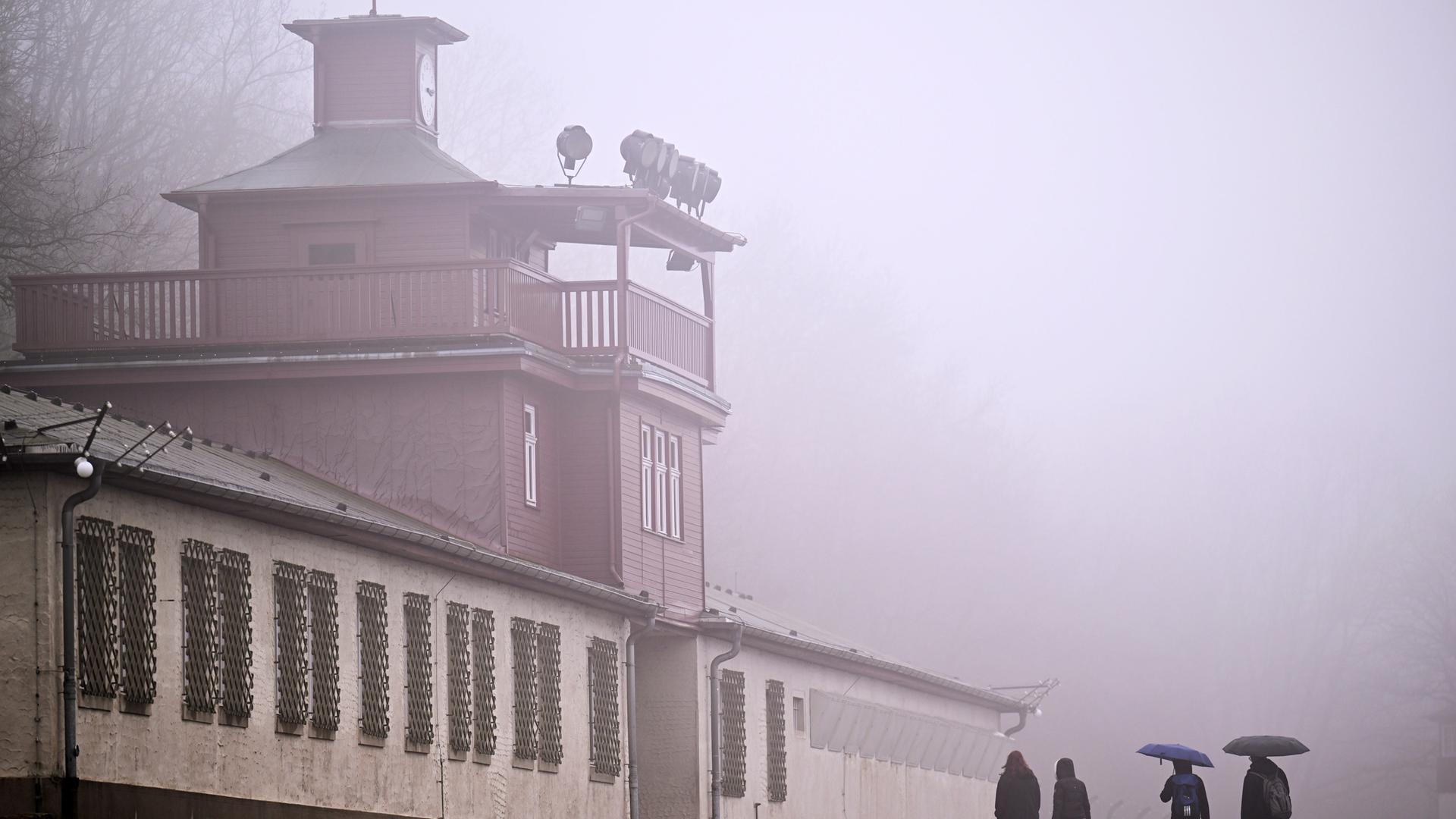 Das Lagertor des ehemaligen KZ Buchenwald. Linker Hand sind Baracken zu sehen, deren Fenster vergittert sind. Es ist ein diesiger Tag, vor dem Tor stehen vereinzelt Menschen mit Regenschirm. 