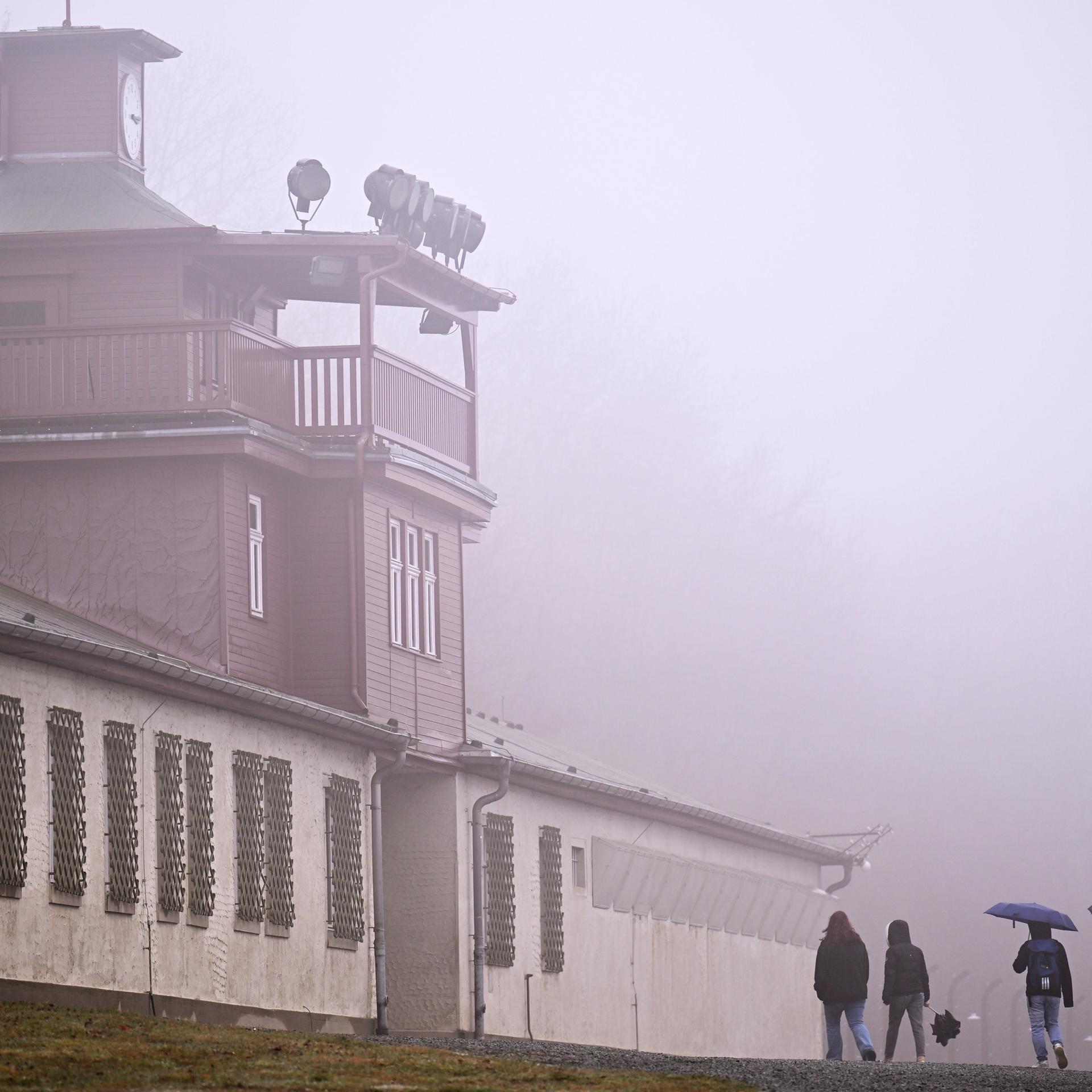 Das Lagertor des ehemaligen KZ Buchenwald. Linker Hand sind Baracken zu sehen, deren Fenster vergittert sind. Es ist ein diesiger Tag, vor dem Tor stehen vereinzelt Menschen mit Regenschirm. 