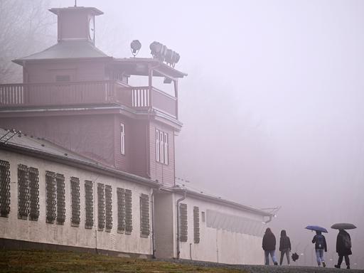 Das Lagertor des ehemaligen KZ Buchenwald. Linker Hand sind Baracken zu sehen, deren Fenster vergittert sind. Es ist ein diesiger Tag, vor dem Tor stehen vereinzelt Menschen mit Regenschirm. 