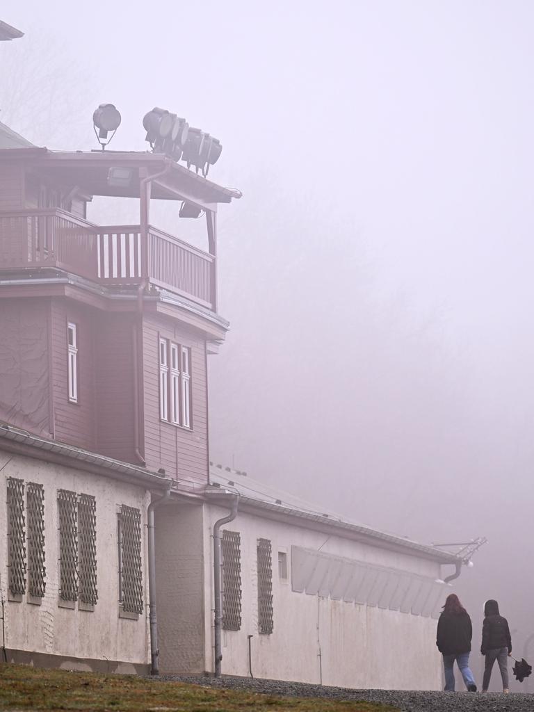 Das Lagertor des ehemaligen KZ Buchenwald. Linker Hand sind Baracken zu sehen, deren Fenster vergittert sind. Es ist ein diesiger Tag, vor dem Tor stehen vereinzelt Menschen mit Regenschirm. 