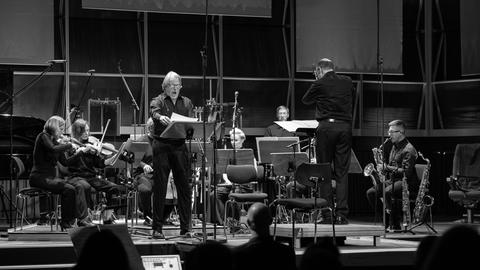 Eion Sänger steht an einem Notenpult und singt, während ihn andere Musiker mit einem Dirigenten auf der Bühne begleiten.