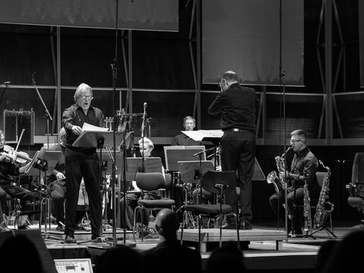 Eion Sänger steht an einem Notenpult und singt, während ihn andere Musiker mit einem Dirigenten auf der Bühne begleiten. Eion Sänger steht an einem Notenpult und singt, während ihn andere Musiker mit einem Dirigenten auf der Bühne begleiten.