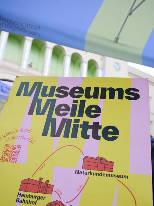 "Museums Meile Mitte" steht auf einem Plakat, im Hintergrund sind Menschen, Sonnenschirme und Stände zu erkennen.