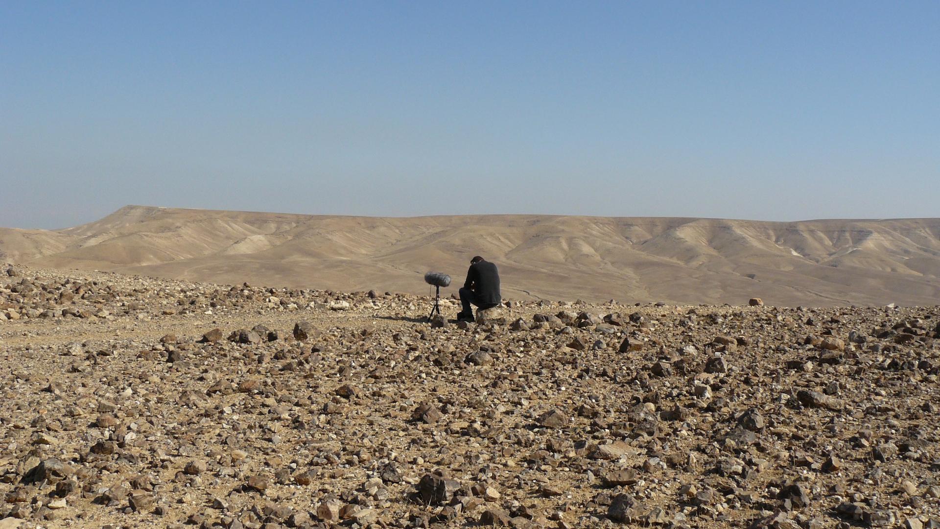 In einer Wüstenlandschaft sitzt der Autor auf einem Stein - neben ihm ein großes Standmikrofon mit Windschutz.