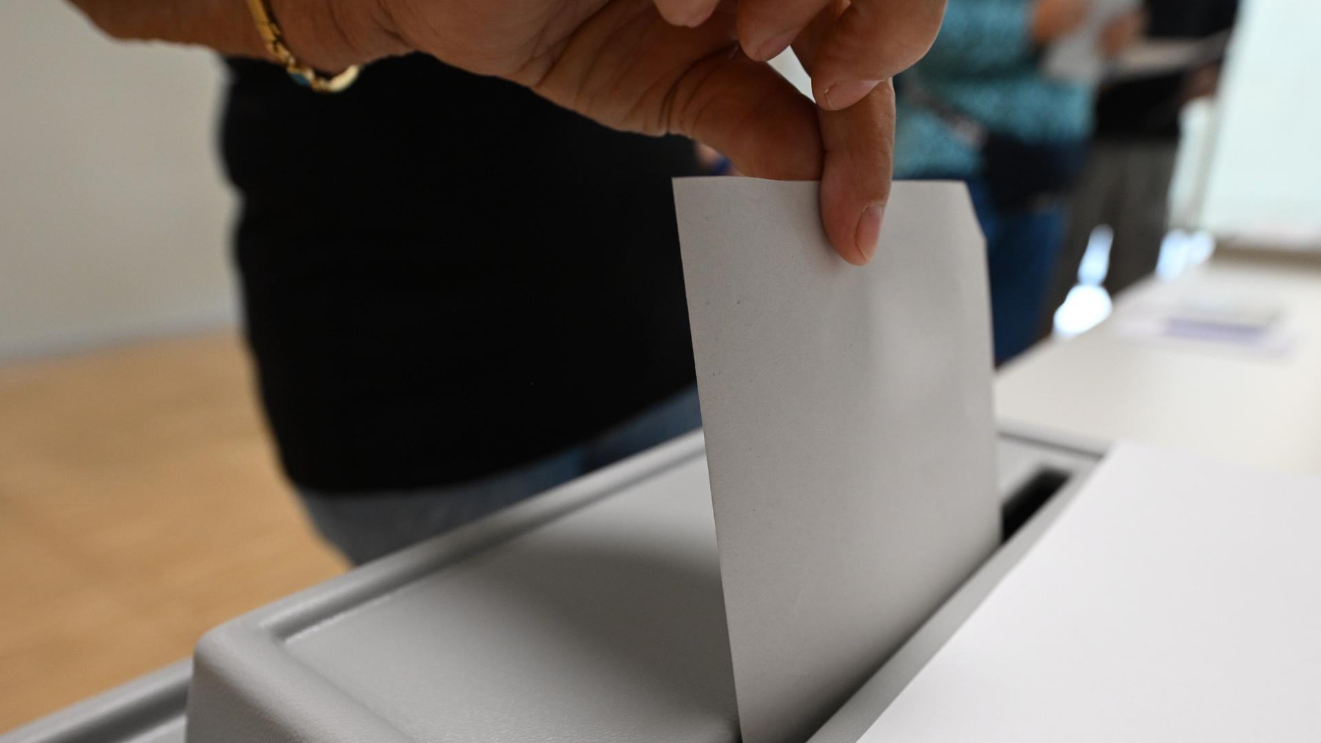 Blick auf eine graue Wahlurne, in die eine Hand ein Wahlzettel steckt. 