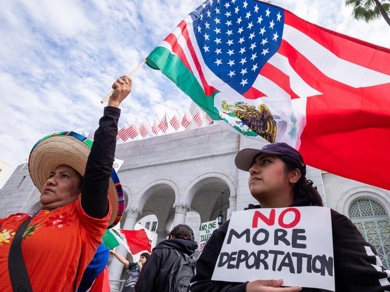 Zwei Demonstrantinnen gegen die Migrationspolitik von Trump in Los Angeles. Eine Frau schwenkt eine US-Fahne, eine trägt ein Schild mit den Worten "No more deportations" (Keine Deportationen mehr) 
