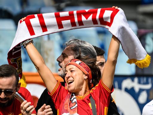 Eine Frau hält einen Schal in die Höhe, auf dem "Ti amo" steht, was auf Italienisch "Ich liebe Dich" bedeutet. Sie schaut glücklich. 