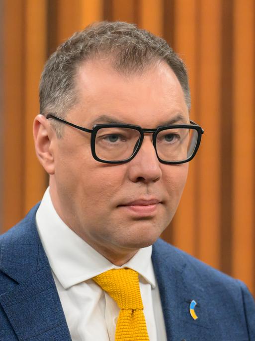 Oleksij Makejew, Botschafter der Ukraine in Deutschland, bei einem Besuch der Talkshow von Caren Miosga. Er trägt einen Anzug und eine dunkle Brille, im Hintergrund ist orange-brauner Stoff zu sehen. 