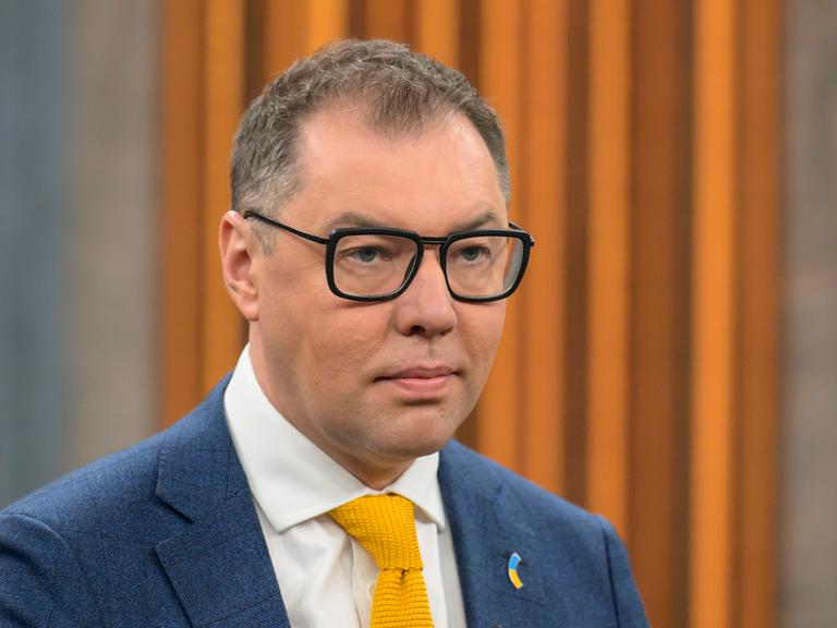 Oleksij Makejew, Botschafter der Ukraine in Deutschland, bei einem Besuch der Talkshow von Caren Miosga. Er trägt einen Anzug und eine dunkle Brille, im Hintergrund ist orange-brauner Stoff zu sehen. 