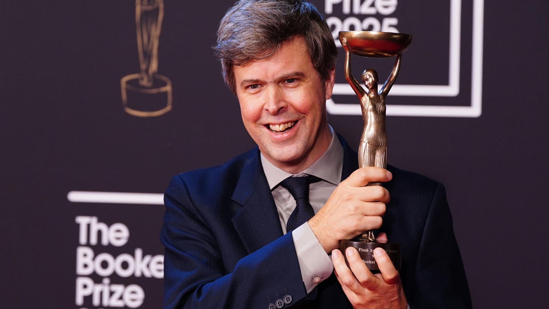Das Foto zeigt den Autor David Szalay, der die Trophäe des Booker Prize in der Hand hält.