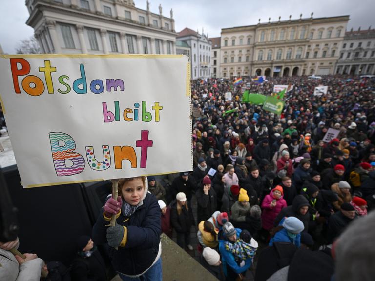 Eine Menschenmenge hat sich auf einem Platz versammelt. Ein Mädchen hält ein Schild in die Höhe. Darauf steht "Potsdam bleibt Bunt". 