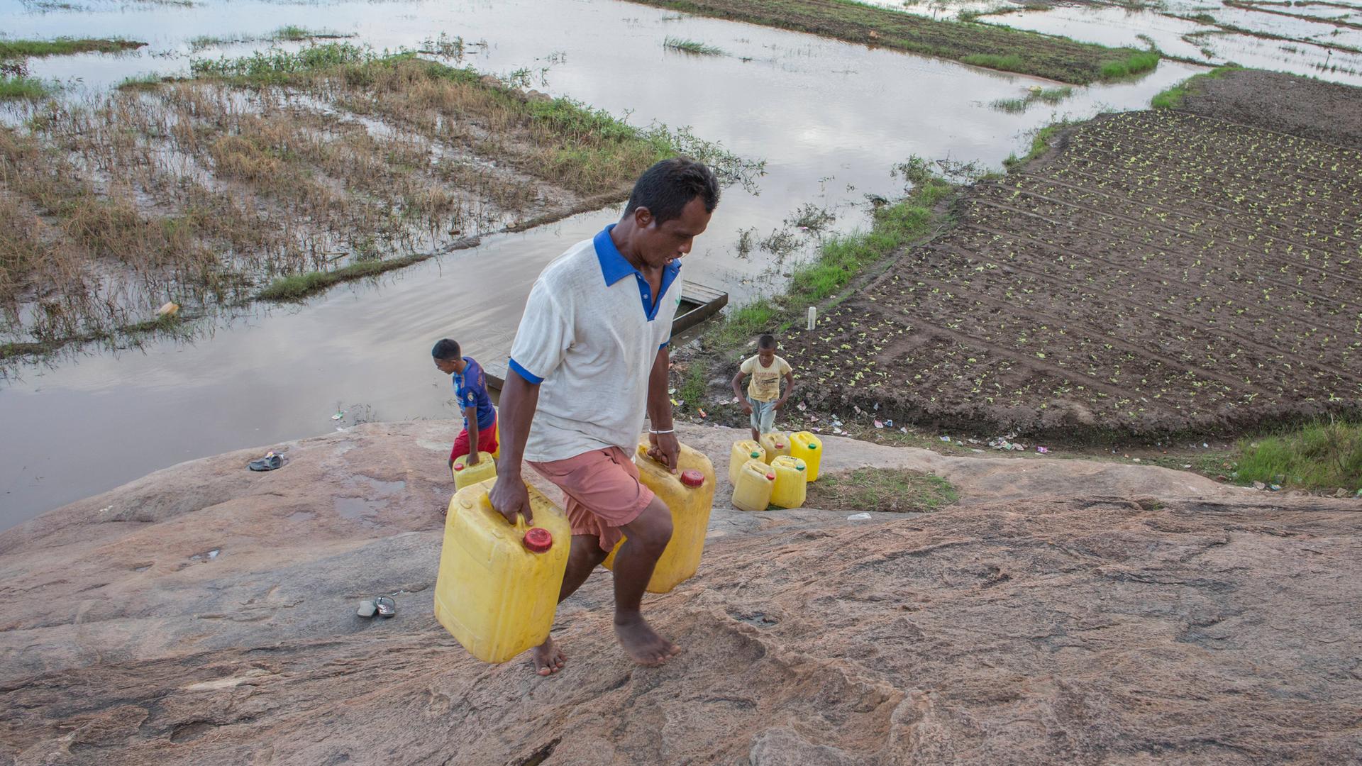 Ein Mann in Madagaskar transportiert Trinkwasser kilometerweit nach Hause. Wasserkanister mit sauberem Trinkwasser werden aus einem 5 Kilometer entfernten Gemeinschaftshahn geholt, der Weg dorthin führt durch Hochwassermassen auf einem Feld
