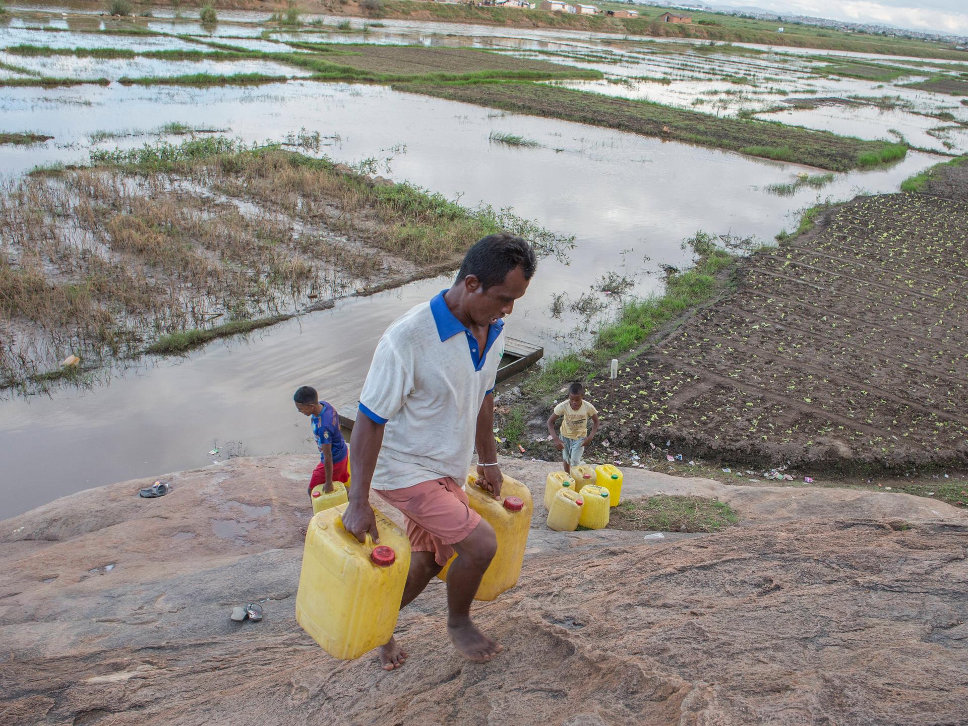 Ein Mann in Madagaskar transportiert Trinkwasser kilometerweit nach Hause. Wasserkanister mit sauberem Trinkwasser werden aus einem 5 Kilometer entfernten Gemeinschaftshahn geholt, der Weg dorthin führt durch Hochwassermassen auf einem Feld