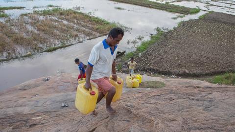Ein Mann in Madagaskar transportiert Trinkwasser kilometerweit nach Hause. Wasserkanister mit sauberem Trinkwasser werden aus einem 5 Kilometer entfernten Gemeinschaftshahn geholt, der Weg dorthin führt durch Hochwassermassen auf einem Feld