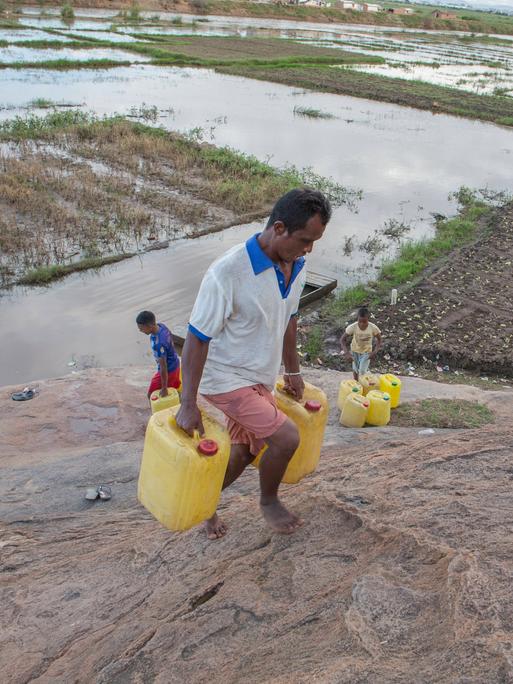 Ein Mann in Madagaskar transportiert Trinkwasser kilometerweit nach Hause. Wasserkanister mit sauberem Trinkwasser werden aus einem 5 Kilometer entfernten Gemeinschaftshahn geholt, der Weg dorthin führt durch Hochwassermassen auf einem Feld