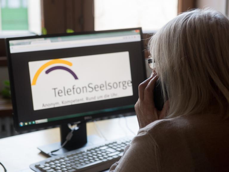 Eine ehrenamtliche Mitarbeiterin der evangelischen Telefonseelsorge München hält einen Telefonhörer an einem Arbeitsplatz für Seelsorger, während die Homepage der Telefonseelsorge auf einem Monitor zu sehen ist.