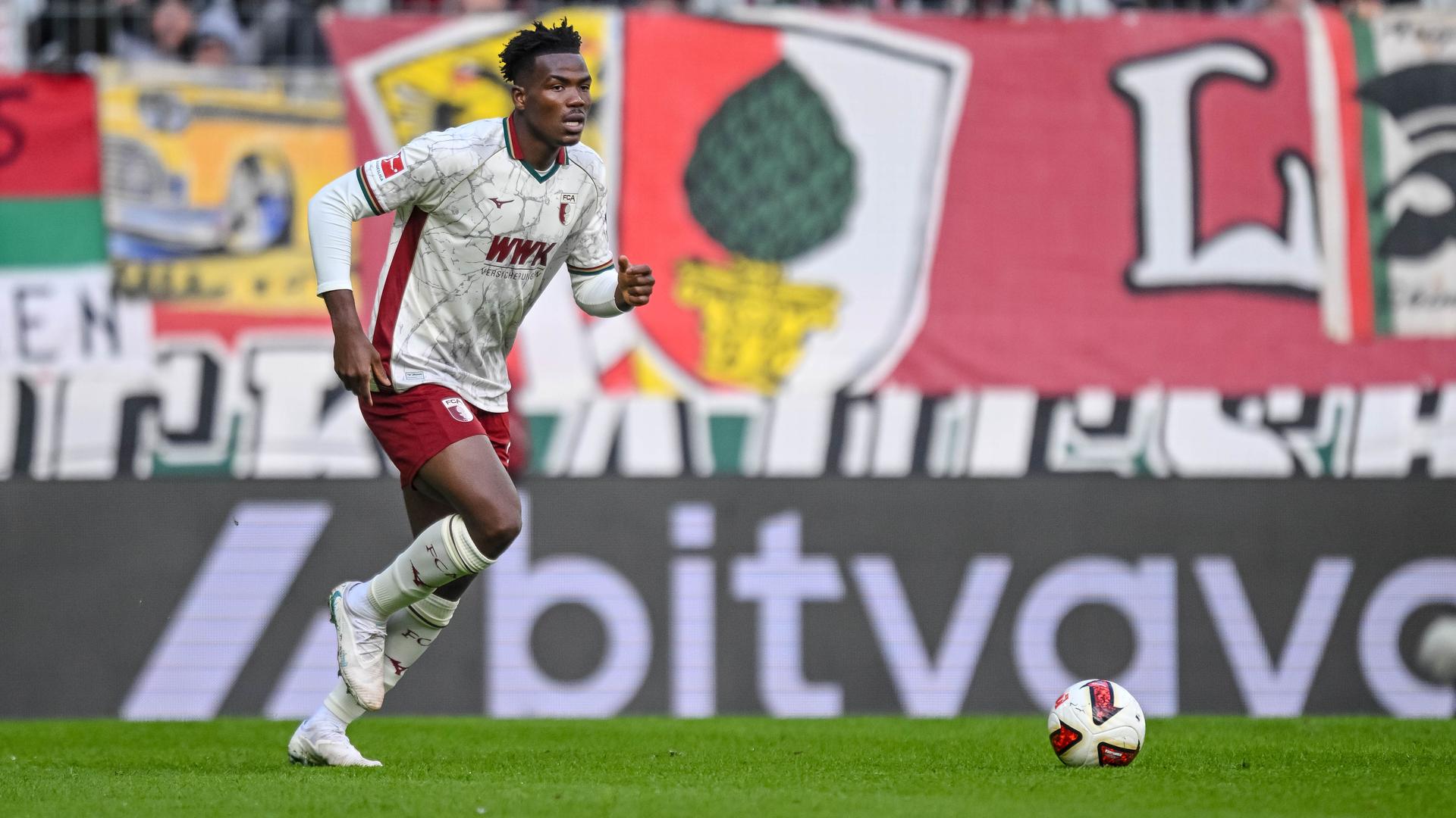 Zu sehen ist der Fußballspieler Juma Bah vom FC Augsburg während eines Bundesligaspiels im Stadion. Er treibt gerade den Ball nach vorne. Dahinter bunte Banner am Trübünenzaun. 