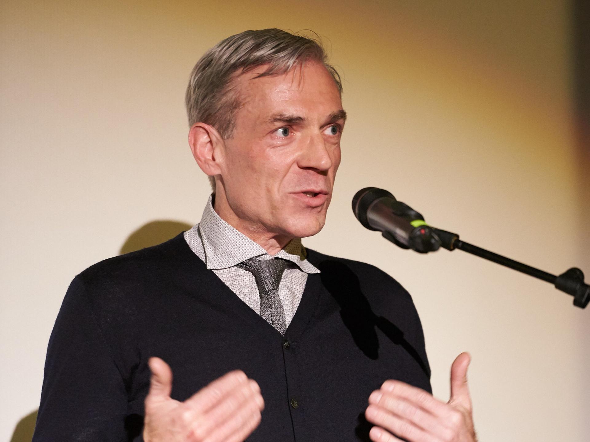 Lars Henrik Gass spricht auf einem Podium in ein Mikrofon. Er trägt kurz geschnittenes graues Haar und einen schwarzen Pullover.