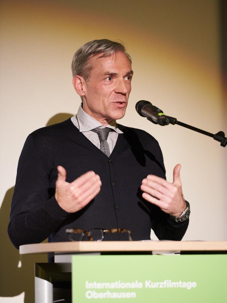 Lars Henrik Gass spricht auf einem Podium in ein Mikrofon. Er trägt kurz geschnittenes graues Haar und einen schwarzen Pullover.