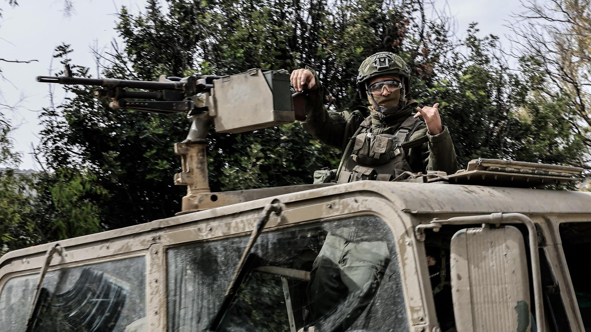Ein israelischer Soldat steht in einem Militärfahrzeug und lehnt einen Arm gegen ein befestigtes Maschinengewehr, während er mit der anderen Hand ein Zeichen gibt. Er trägt Helm und Schutzbrille.