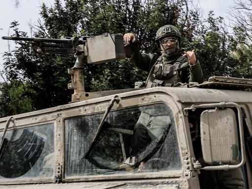 Ein israelischer Soldat steht in einem Militärfahrzeug und lehnt einen Arm gegen ein befestigtes Maschinengewehr, während er mit der anderen Hand ein Zeichen gibt. Er trägt Helm und Schutzbrille.