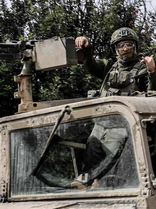Ein israelischer Soldat steht in einem Militärfahrzeug und lehnt einen Arm gegen ein befestigtes Maschinengewehr, während er mit der anderen Hand ein Zeichen gibt. Er trägt Helm und Schutzbrille. Ein israelischer Soldat steht in einem Militärfahrzeug und lehnt einen Arm gegen ein befestigtes Maschinengewehr, während er mit der anderen Hand ein Zeichen gibt. Er trägt Helm und Schutzbrille.