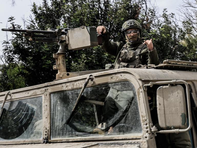 Ein israelischer Soldat steht in einem Militärfahrzeug und lehnt einen Arm gegen ein befestigtes Maschinengewehr, während er mit der anderen Hand ein Zeichen gibt. Er trägt Helm und Schutzbrille. Ein israelischer Soldat steht in einem Militärfahrzeug und lehnt einen Arm gegen ein befestigtes Maschinengewehr, während er mit der anderen Hand ein Zeichen gibt. Er trägt Helm und Schutzbrille.