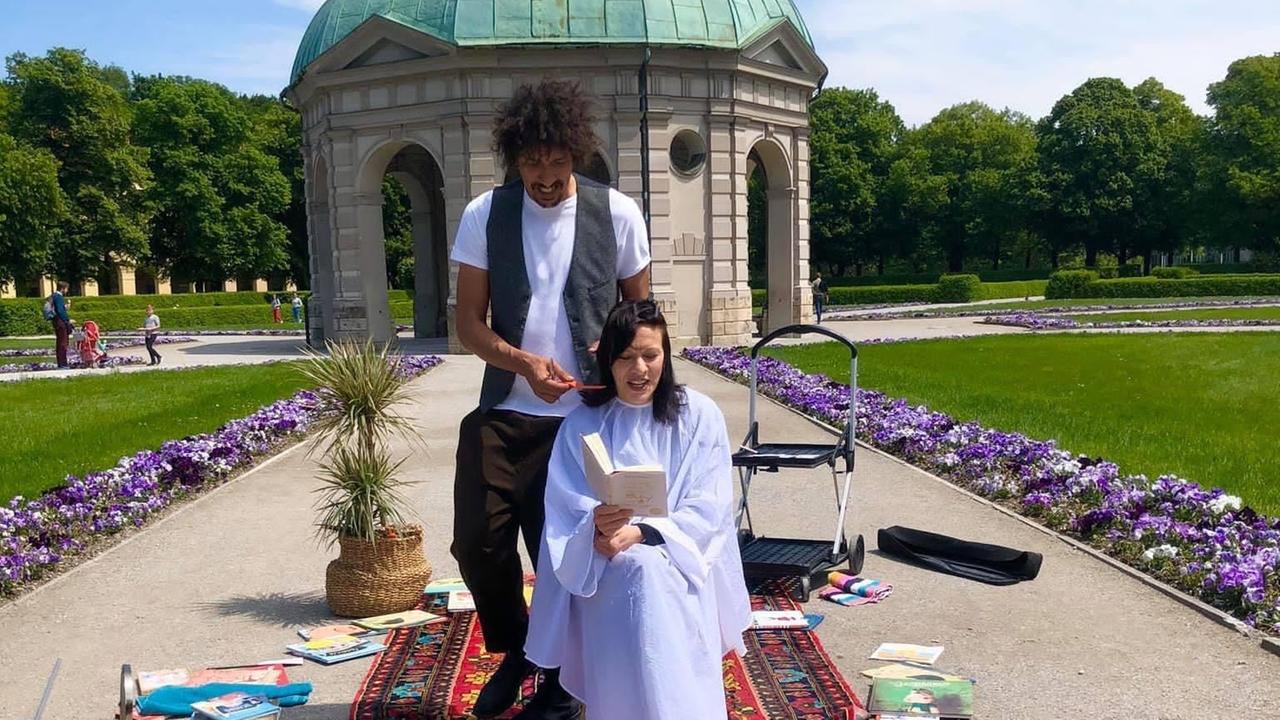 Ein Friseur schneidet einer Frau in einem Park die dunklen Haare. Sie liest dabei in einem Buch.