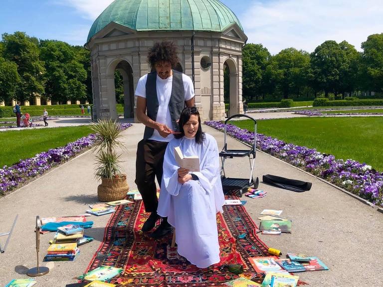 Ein Friseur schneidet einer Frau in einem Park die dunklen Haare. Sie liest dabei in einem Buch.