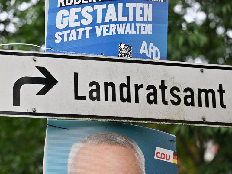 Plakate mit Fotos der Kandidaten hängen neben einem Wegweiser zum Landratsamt. 