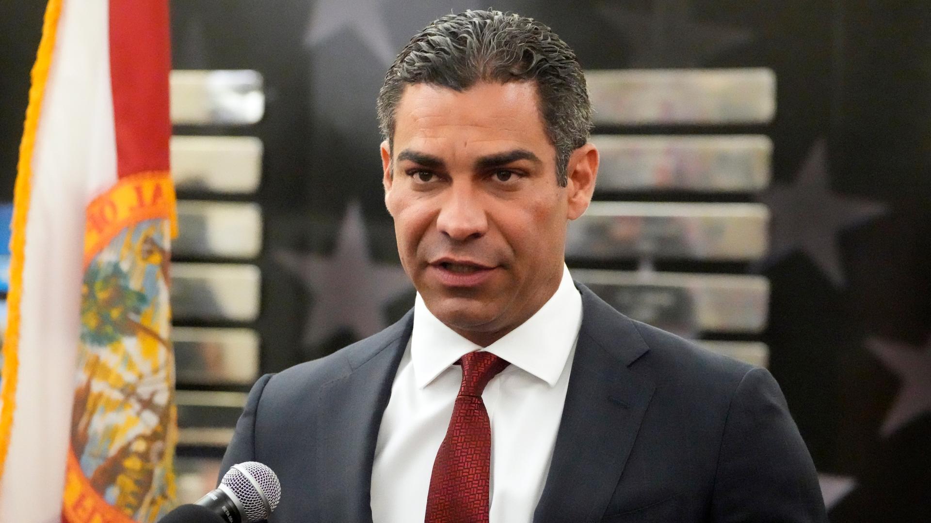 Suarez im dunklen Anzug, mit weinroter Krawatte und mit zurückgegelten Haaren steht vor einer Wand und sagt etwas in ein Mikrofon. Daneben die Flagge Floridas. Suarez im dunklen Anzug, mit weinroter Krawatte und mit zurückgegelten Haaren steht vor einer Wand und sagt etwas in ein Mikrofon. Daneben die Flagge Floridas.