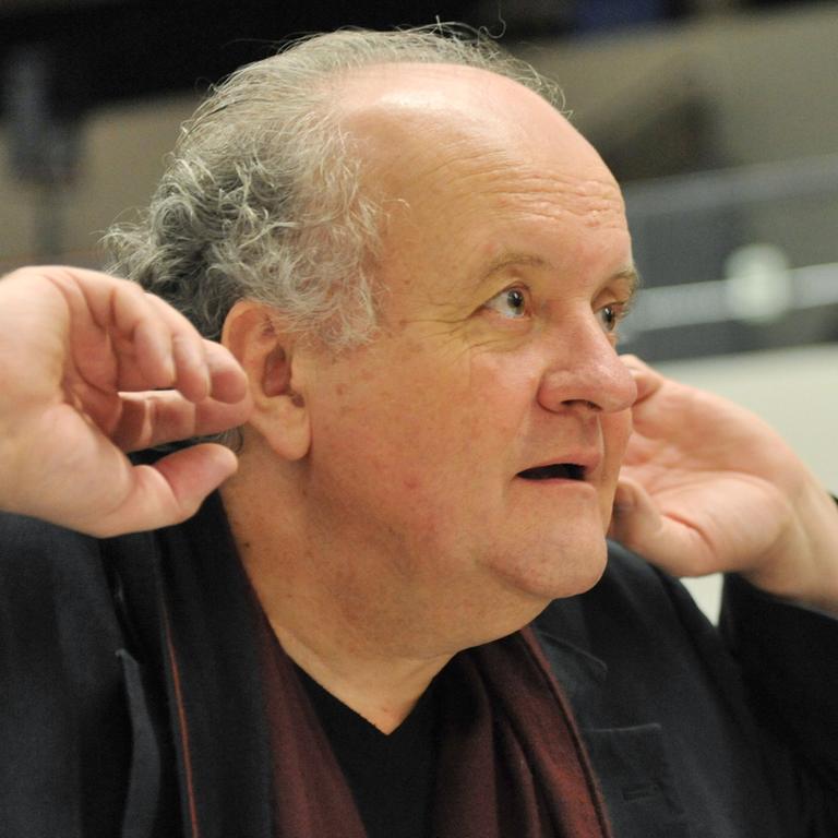 Ein älterer Mann mit schütterem Haar blickt aufmerksam. Er hält seine Hände an die Ohren, um besser zu hören. Es ist der Komponist Wolfgang Rihm.
