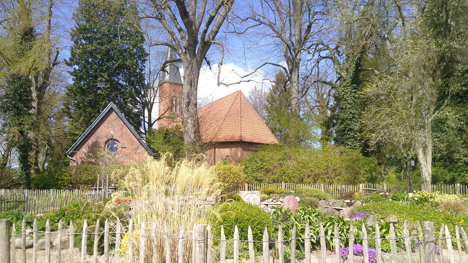 Im Vordergrund ist ein "liturgischer Garten" zu sehen, im Hintergrund, von Bäumen umgeben, die Kirche und ein Gemeindegebäude aus rotem Backstein. Im Vordergrund ist ein "liturgischer Garten" zu sehen, im Hintergrund, von Bäumen umgeben, die Kirche und ein Gemeindegebäude aus rotem Backstein.