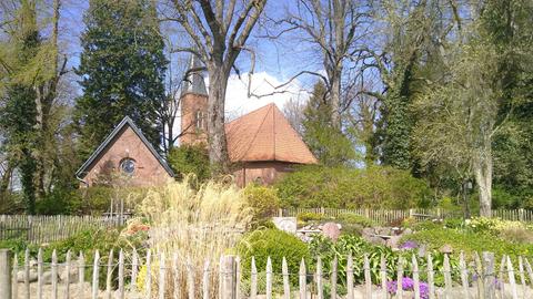 Im Vordergrund ist ein "liturgischer Garten" zu sehen, im Hintergrund, von Bäumen umgeben, die Kirche und ein Gemeindegebäude aus rotem Backstein.
