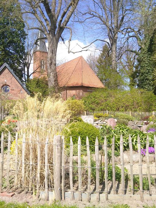 Im Vordergrund ist ein "liturgischer Garten" zu sehen, im Hintergrund, von Bäumen umgeben, die Kirche und ein Gemeindegebäude aus rotem Backstein.