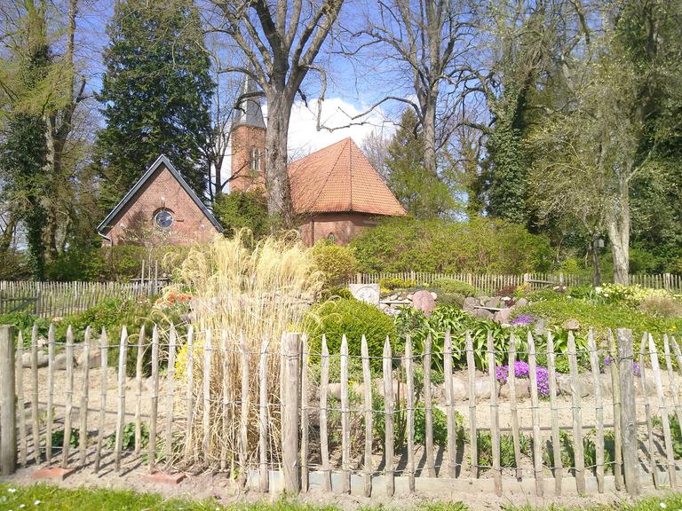 Im Vordergrund ist ein "liturgischer Garten" zu sehen, im Hintergrund, von Bäumen umgeben, die Kirche und ein Gemeindegebäude aus rotem Backstein.